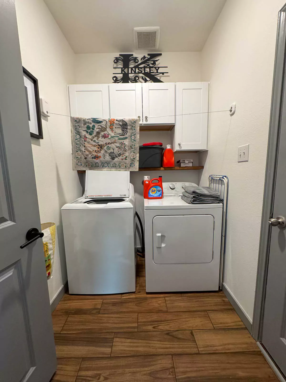 Finished DIY makeover of a small laundry room featuring stacked washer and dryer, floating wood shelves, white cabinetry, and silver hardware. Clean, modern design with efficient use of vertical space and warm, minimal styling.